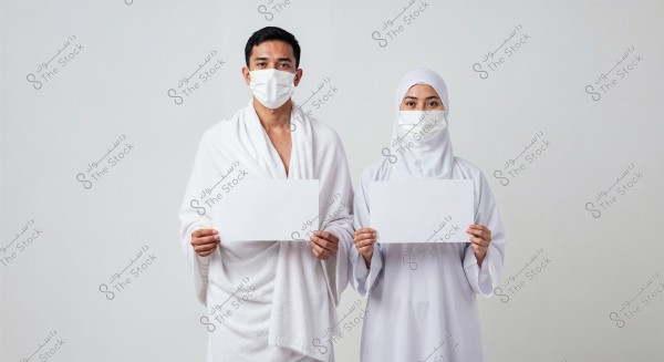 A portrait of a man and a woman standing against a white background. The man is wearing a white Ihram and a white medical mask, holding a blank white piece of paper. The woman is wearing a white hijab and abaya, also with a medical mask, and holding a blank white sheet of paper. The setting suggests a context related to Hajj or Umrah.