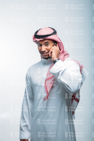 رجل سعودي يرتدي  الزي الرسمي السعودي ، على خلفية بيضاء ، يتحدث مبتسماً عبر الجوال ، مكالمات الأهل والأصدقاء ، لحظة سعيدة