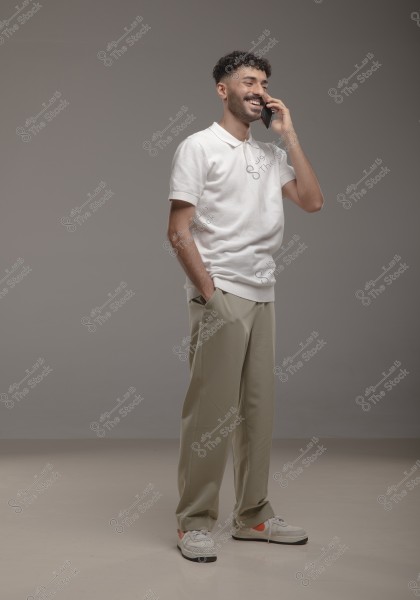 A portrait of a man standing, smiling, and talking on the phone in a studio with a gray background. He is wearing a white polo shirt, beige trousers, and white sneakers. His appearance is modern and stylish.