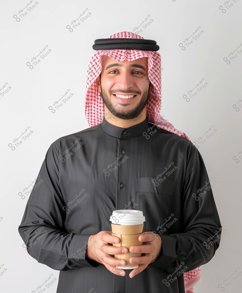 Portrait of a smiling man wearing traditional Saudi attire, including a black thobe, red and white checkered ghutra, and an agal. He is holding a paper coffee cup with both hands. The white background enhances the details of the traditional clothing.