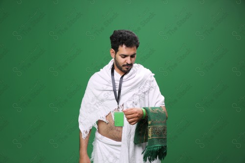 Portrait of a Saudi pilgrim on a green background wearing the Ihram to perform Hajj, holding a carpet in his hand in several positions