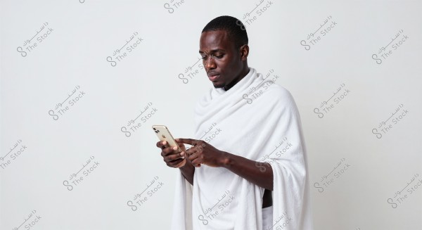يظهر في الصورة رجل يرتدي إحرامًا أبيض اللون, وهو اللباس التقليدي الذي يُرتدى أثناء أداء مناسك الحج أو العمرة. الرجل يقف أمام خلفية بيضاء, ينظر إلى هاتف ذكي بيده ويبدو مركزًا في ما يراه.