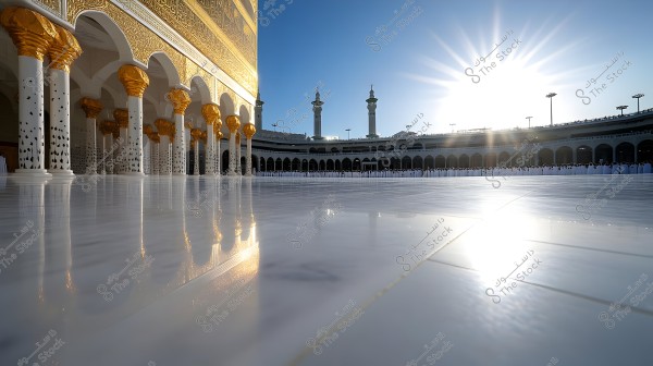 An image showing the holy site in Mecca, featuring a section of the Kaaba and part of the surrounding courtyard. The golden, patterned columns are part of the stunning architecture, with bright sunlight reflecting off the shiny white flooring. The sky is clear blue, and the historic minarets of the sanctuary are visible in the background.