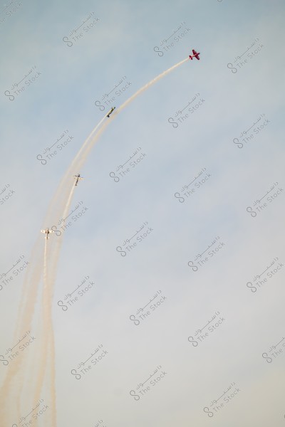 Four stunt planes flying in a clear blue sky, leaving long smoke trails forming an arc in the air. A red plane is leading, followed by three other white or silver planes.