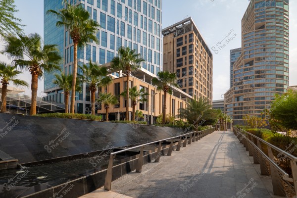 Urban scene showcasing modern high-rise buildings with glass facades surrounded by green palm trees. In the foreground, there is a paved walkway with metal railings and a water feature with black panels cascading water, surrounded by greenery. The buildings exhibit varied architectural designs and intricate geometric details.