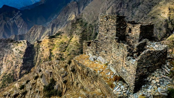 The image shows a natural landscape of a mountain in the Asir region, Saudi Arabia, featuring an old stone building positioned beside steep rocky slopes and green terraces. The building appears to be part of a traditional village amidst a stunning mountainous environment that stretches out to the distant horizon.
