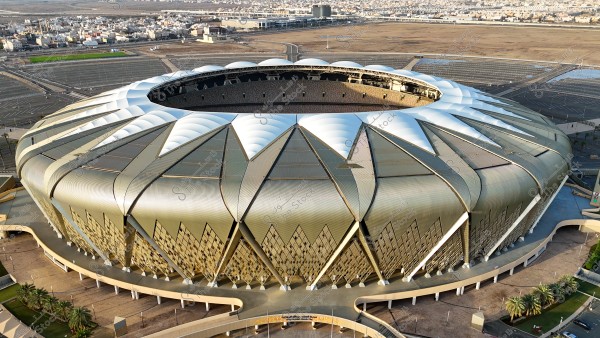 صورة لملعب رياضي دائري كبير في مدينة جدة, المملكة العربية السعودية. يتميز بتصميم سقف حديث يضم أشكالاً هندسية ذهبية وفضية. تُحاط الملعب بمناظر طبيعية وأشجار نخيل, بينما تظهر في الخلفية مساحات واسعة وعدد من المباني السكنية.