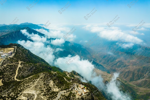 An aerial view of a mountain range covered in clouds in the distance. The steep mountainous terrain is dotted with trees and vegetation, with a winding road along the mountains. A few buildings are visible in the bottom right corner. The sky is clear with some white clouds surrounding the mountains.