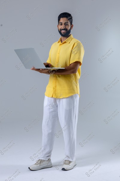 An image of a man standing and smiling while holding a laptop. The man is wearing a yellow short-sleeved shirt, white pants, and white sneakers. The background is grey.