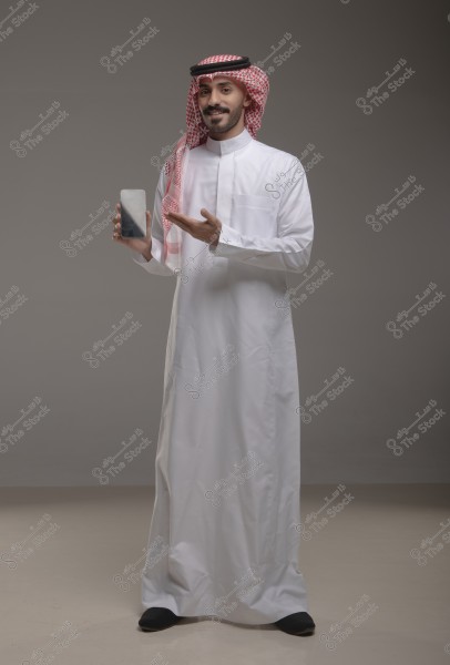 صورة لرجل يرتدي الثوب التقليدي السعودي والغترة الحمراء والعقال الأسود, يقف في استوديو رمادي ويعرض هاتفًا ذكيًا بيده. تبدو ملامحه مبتسمة وثقته بالنفس واضحة.
