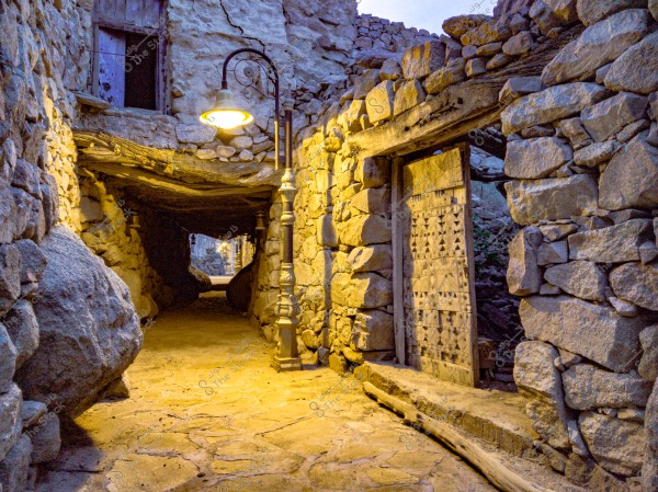 An old stone passageway with large stone walls and a long archway, illuminated by warm yellow lights. A large wooden door with carved geometric patterns is visible on the right side.