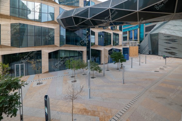 A photograph of a modern courtyard surrounded by glass buildings with a unique geometric design. The glass facade creates a futuristic pattern with clear lines and multiple faceted protrusions. Walkways stretch across the neatly arranged pavement. A few trees are scattered around the courtyard, along with tall light posts, and a glass bridge extends between the buildings.