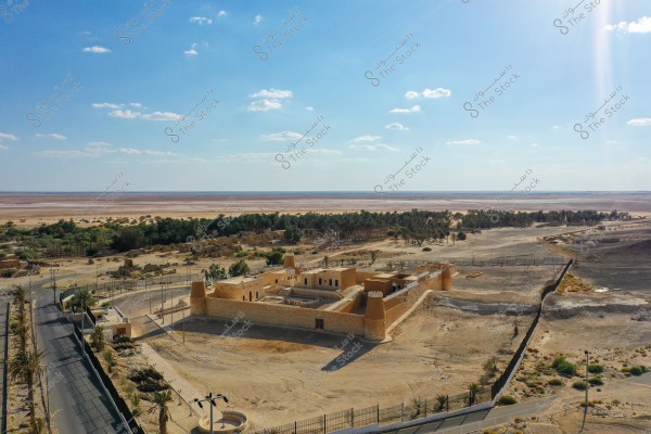 منظر جوي لموقع أثري في منطقة صحراوية, يظهر فيها قلعة قديمة محاطة بجدران مرتفعة وأبراج. يجاور القلعة طريق محاط بأشجار نخيل, ويمتد خلفها واحة بها مجموعة من الأشجار. السماء زرقاء صافية مع بعض السحب الصغيرة.