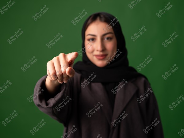 Image of a woman wearing a black hijab, extending her hand forward with a finger pointing. The background is green, and the woman is wearing a dark-colored coat.