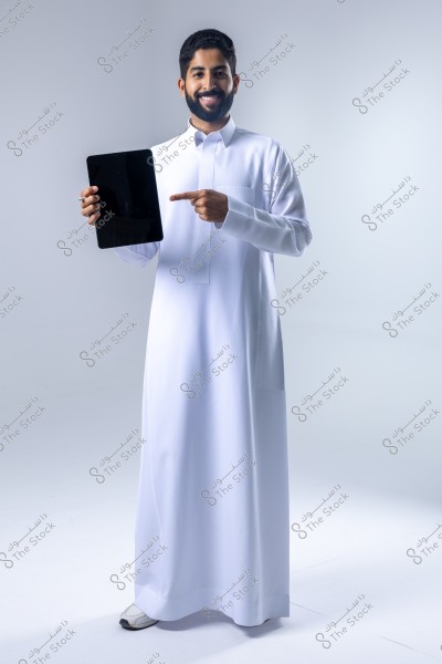 An image of a man wearing a white thobe, which is a traditional garment suggesting a Saudi origin. The man is standing and smiling in a studio with a white background, holding a tablet in his left hand and pointing at it with his right finger.