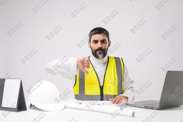 A man sits at a desk wearing a work outfit consisting of a white shirt and a yellow safety vest with reflective strips. His expression is serious, and he gestures with a thumbs-down. In front of him on the desk is a white helmet, alongside a laptop, and several papers and plans. The overall clothing style is possibly linked to the Middle East.