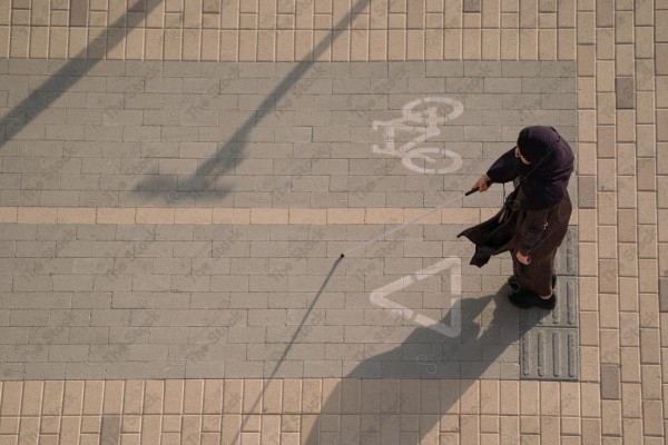 امرأة "من ذوي الاعاقة" تستخدم عصا بيضاء للمشي على رصيف مع مسار مرسوم للدراجات الهوائية.