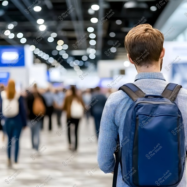 A man wearing a light-colored shirt and carrying a blue backpack stands in a crowded venue resembling a conference hall. The background features many people walking under bright lighting and a ceiling with hanging fixtures.