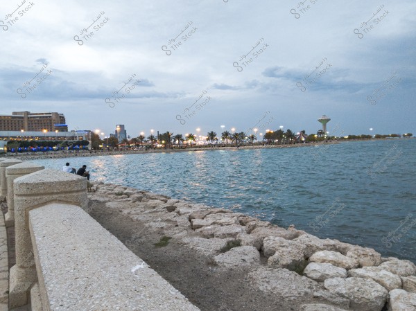 An image showing a scenic view of a seafront in Dammam, Saudi Arabia. There is a stone pier on the left extending along the coast, with two people sitting on it. The blue water stretches to the horizon where a city is lit up with evening lights. Palm trees are visible along the promenade, and a distinctive water tower is seen on the right side of the image.