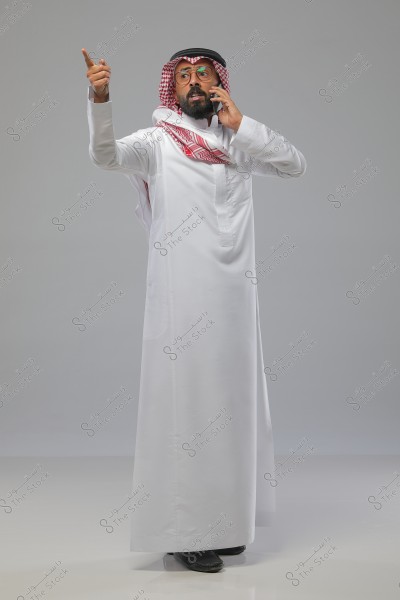 A portrait of a man standing in a studio, wearing a white traditional thobe, a red checkered headscarf, and a black agal, suggesting he is from the Gulf region. He is wearing glasses and talking on a mobile phone while pointing upwards with his finger. The background is a neutral gray.