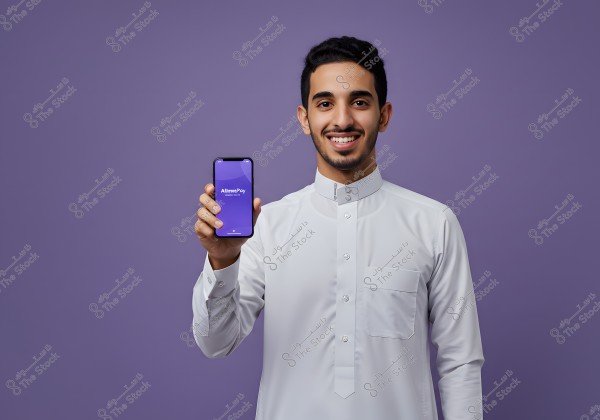 Image of a young man wearing a traditional white thobe, smiling against a light purple background. The man is holding a mobile phone displaying the \"AlmaraPay\" app. The clothing appears to reflect a Saudi style.