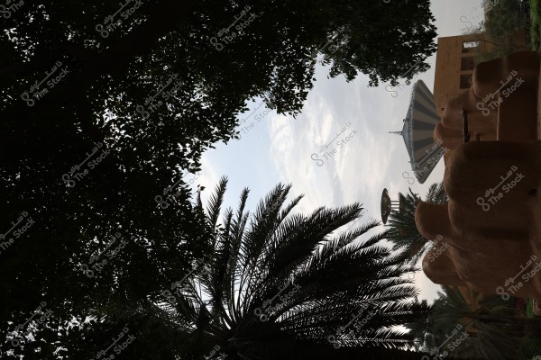 The image depicts a natural landscape featuring tall palm trees, with traditional mud buildings in the foreground. In the background, there is a large cone-shaped building with colored stripes, surrounded by green trees. Light filters through the branches, creating an aesthetic view against a slightly cloudy blue sky.