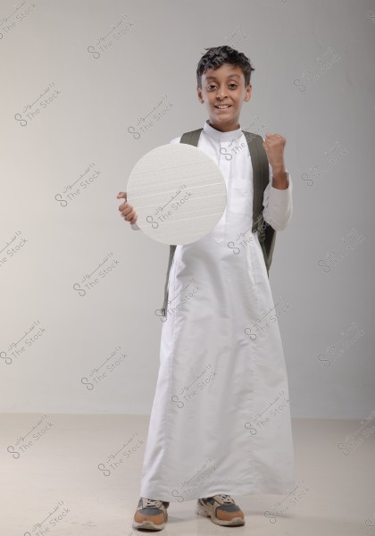Image of a boy wearing a traditional white thobe and carrying a backpack. He is smiling at the camera while holding a large white circle in his right hand and raising his left fist in a gesture of triumph or celebration. The floor and background wall are plain and light-colored.