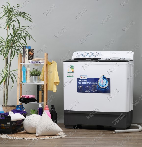 The image shows a large white washing machine with a \"DISON\" brand label on the right side. Behind the washing machine, there is a grey wall and a brown wooden floor. On the left, there is a wooden shelf unit holding various items, including folded clothes, a hanging yellow shirt, houseplants, and detergent bottles. In front of the washing machine, there is a laundry basket with colorful clothes and round comfy pillows on a small rug.