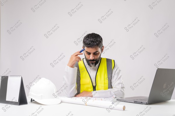 An image of a middle-aged man sitting at a desk wearing a yellow safety vest. The man appears focused while looking at a large blueprint in front of him. Beside the blueprint, there is a white safety helmet and a laptop. The background is completely white, highlighting the man and the desk.