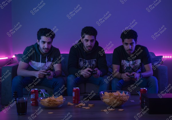 Three men sitting on a sofa in a room lit with purple neon lights, holding video game controllers and focused on playing. They are wearing casual, trendy clothing, including shirts in various colors and jeans. There are bowls of potato chips and cans of soda on the table in front of them.