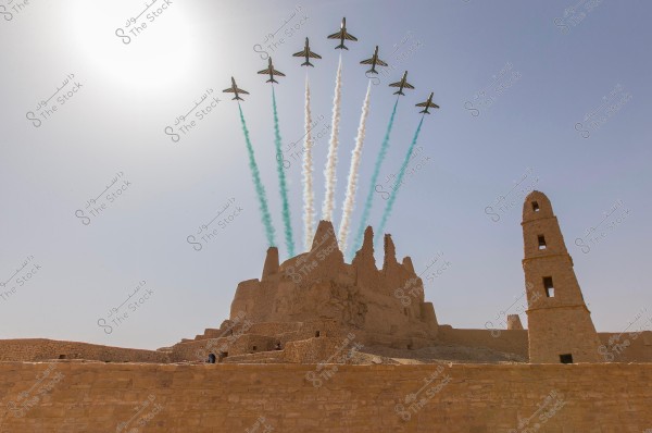 Aircraft performing an air show over historical mudbrick ruins in Saudi Arabia. The planes fly in a coordinated formation, leaving trails of white and green smoke in the blue sky. The sun is prominently visible in the background, and the mudbrick ruins include towers and crumbling walls.