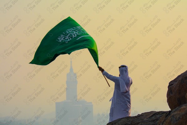 A snapshot of a Saudi man in traditional Saudi dress holding the Saudi flag over a mountain in the Makkah Al-Mukarramah region, the sky, the royal clock tower building in the Grand Mosque, buildings and landmarks, the Grand Mosque of Mecca.