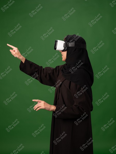 امرأة ترتدي حجاب ونظارة واقع افتراضي, وتشير بيدها إلى الأمام بخلفية خضراء.
