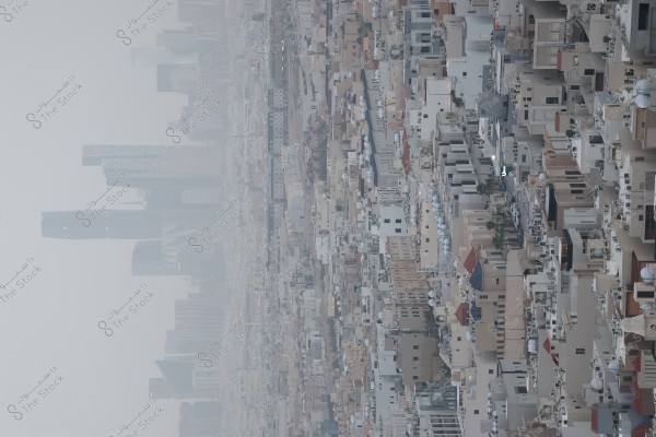 An image of a modern city featuring a cluster of closely-packed residential buildings with diverse architectural designs in beige and white colors. In the background, a haze partially obscures some tall skyscrapers, giving the skyline a slightly blurred appearance. The city appears uncrowded, with a few trees lining the side streets.