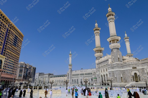 Image showing the exterior courtyard of the Grand Mosque in Mecca, Saudi Arabia, highlighting the magnificent minarets and surrounding architectural structures. The image features numerous pilgrims and worshippers wearing white Ihram garments and other traditional clothing, walking in the courtyard under a clear blue sky.