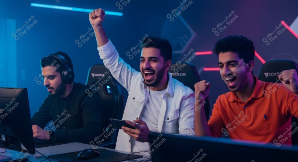 Image of three men playing video games in a room with blue and pink neon lighting. The person on the left is wearing headphones and a black shirt, focused on a computer screen. The person in the middle is wearing a white shirt, raising his hand in celebration and holding a mobile phone. The person on the right is wearing a polo shirt, cheering enthusiastically. The atmosphere reflects excitement and competition in an intense gaming environment.