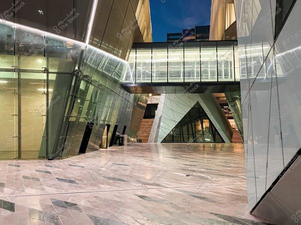 An image of a modern architectural building featuring angled reflective glass walls. There is an overhead glass walkway connecting parts of the building. The floor is covered with multicolored marble tiles. The artificial lighting adds an elegant touch to the area in the evening.