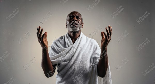 An image of an elderly man wearing a white garment, resembling a Hajj ihram, with his hands raised as if in a position of prayer or meditation. His facial expression is serious and contemplative. The background is a simple gray color.