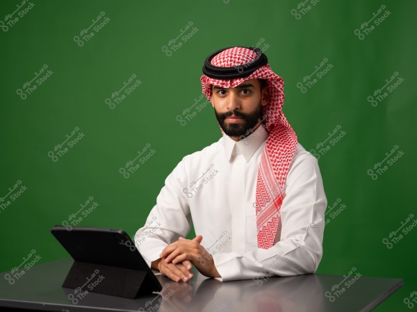 A man sitting in front of a tablet, wearing traditional Saudi attire, with a green background.