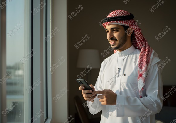 Portrait of a man wearing a white Saudi thobe and a red shemagh standing by a window. He is smiling while looking at a mobile phone in his hand. Indoor lighting and a table lamp are visible in the background.
