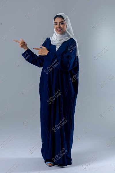 A portrait of a woman wearing a dark blue abaya and a decorative white hijab. She is smiling and pointing both hands to the left. The background is plain gray.