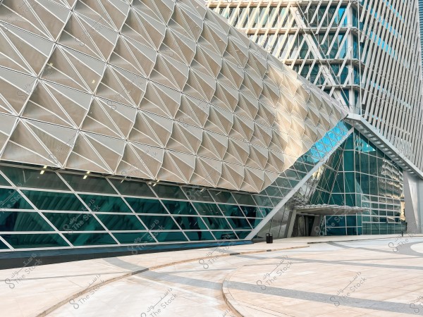 The image shows a modern building with a glass facade characterized by a complex architectural design featuring triangular and rectangular shapes. The glass elements combine with light shadows, giving the building a futuristic appearance. The ground in front of the building is tiled and contains subtle color gradients.