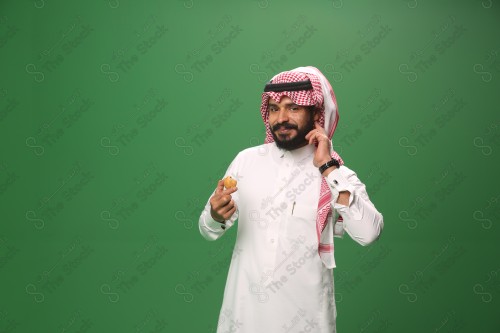 شاب سعودي على خلفية خضراء يرتدي الثوب و الشماغ، يقوم بعمل حركات بيده، يأكل الكعكة ، احتفالات العيد