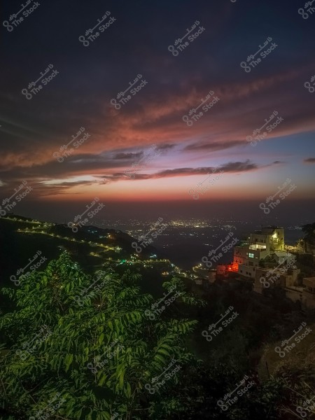 صورة تظهر منظرًا طبيعيًا خلال الغروب, حيث تملأ الألوان البرتقالية والبنفسجية السماء وتنعكس على السحب. تظهر في الأسفل التلال التي تحيط بها إضاءة المنازل, وبعض الأشجار الخضراء في المقدمة. تظهر أيضًا مدينة بعيدة بإنارتها الليلة.