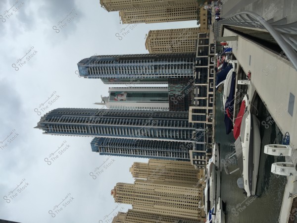 Image of Dubai Marina featuring a collection of modern skyscrapers situated along the canal. There are also yachts and small boats docked at the pier beside the water. The sky is overcast, suggesting cloudy weather.