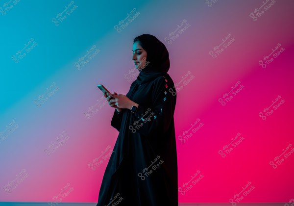 Portrait of a woman wearing a black abaya and hijab, standing sideways while using a mobile phone in her hand. The background features a floor and wall illuminated with multicolored lighting, including blue and pink, creating a modern and distinctive atmosphere.
