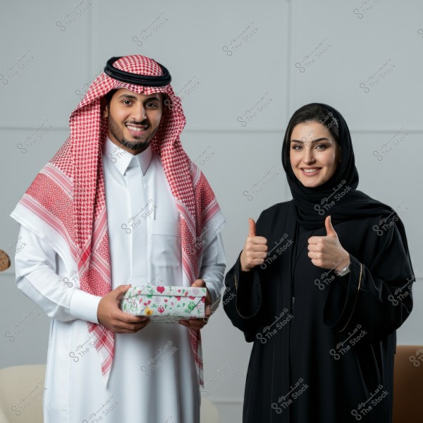The image shows a man and a woman standing together, smiling. The man is wearing traditional Saudi attire, a white thobe, and a red shemagh, holding a gift box with colorful decorations. The woman is wearing a black abaya and hijab, giving a thumbs-up. Both appear happy in a neutral indoor setting.