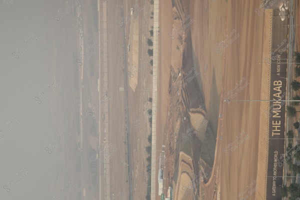 Image of a large construction site showing vast areas of excavated land prepared for building. There is a wall running along the land and some scattered trees. A slogan in English, \"THE MUKAAB,\" is visible on the right, with \"A GATEWAY TO ANOTHER WORLD\" beneath it. The primary colors in the image are various shades of brown and earth tones.
