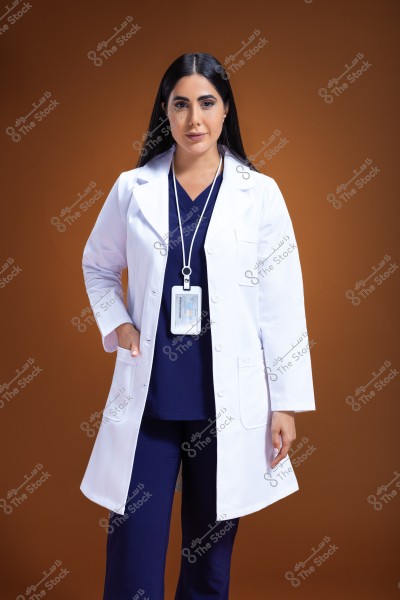 A portrait of a female doctor wearing a white coat and blue medical scrubs, with an identification badge hanging around her neck. She stands with a subtle smile against a solid brown background.
