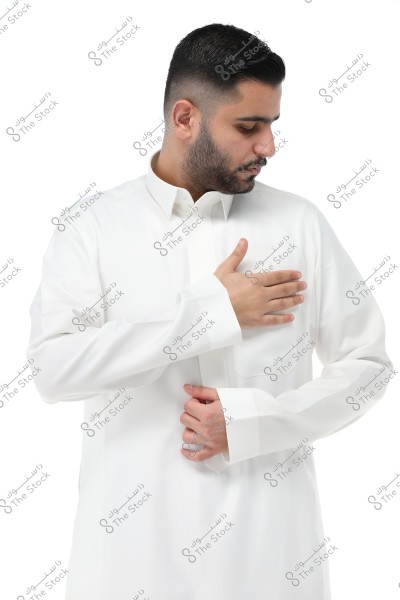 A portrait of a man wearing a traditional white thobe. The man is looking to the right and holding his thobe with his right hand. His hair is short and neatly styled.